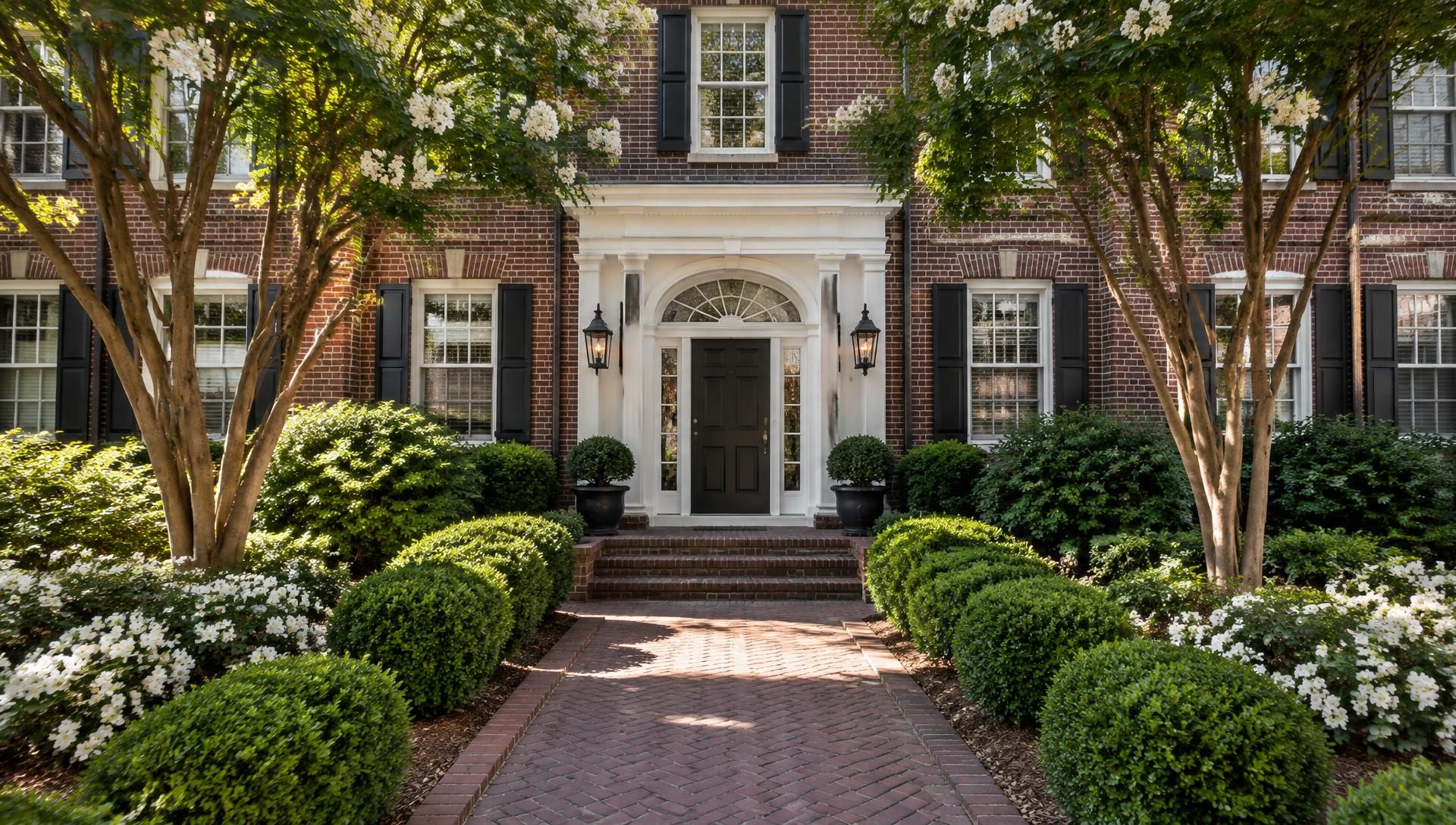 A herringbone brick walkway between clipped boxwoods and flowering crepe myrtles leading to the white-trimmed entrance of a brick apartment building in morning light